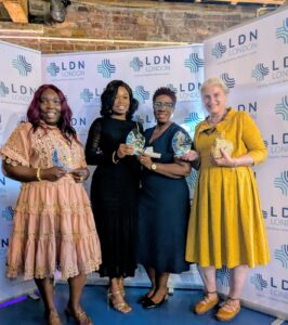 Four women stand proudly in front of an LDN London banner, smiling and holding glass awards. They are dressed in elegant attire, celebrating their achievements.